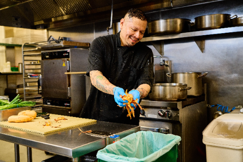 A chef in a commercial kitchen peeling sweet potatoes and placing the peels into a green organics recycling bin.