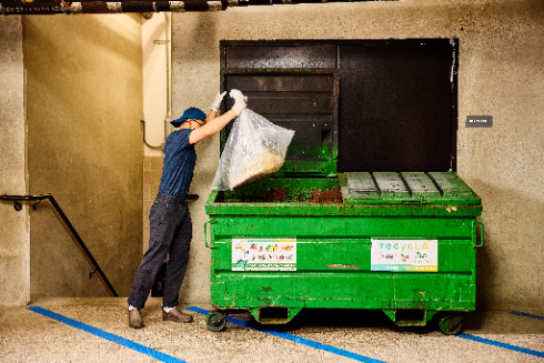 A worker outdoors emptying a bag of organic waste from a small green collection bin into a large green industrial organics dumpster.