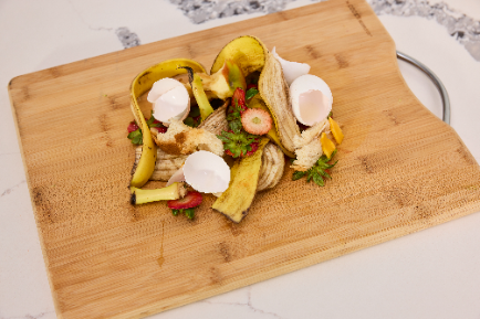 Various food scraps including banana peels, eggshells, and strawberry tops piled on a wooden cutting board.