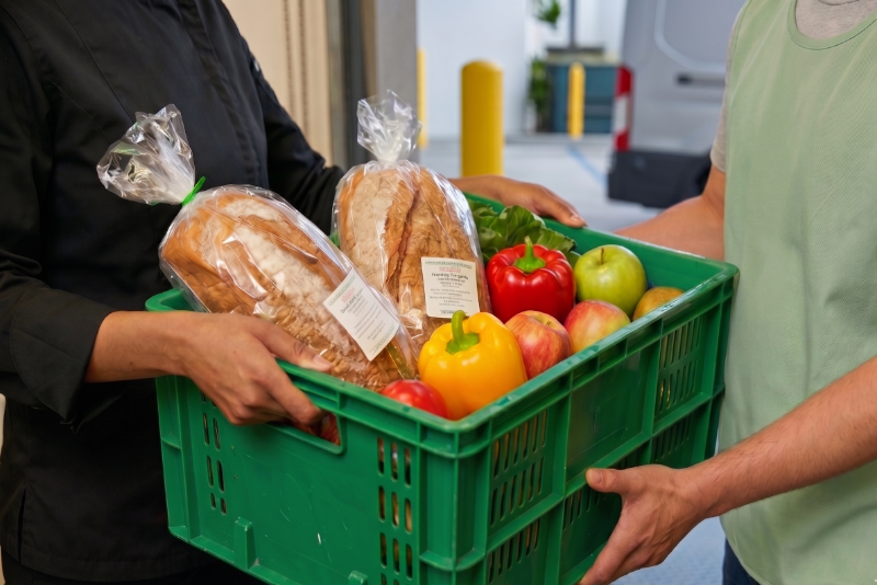 A kitchen staff member in a black uniform hands a green crate filled with fresh bread and produce to a person wearing a green safety vest.