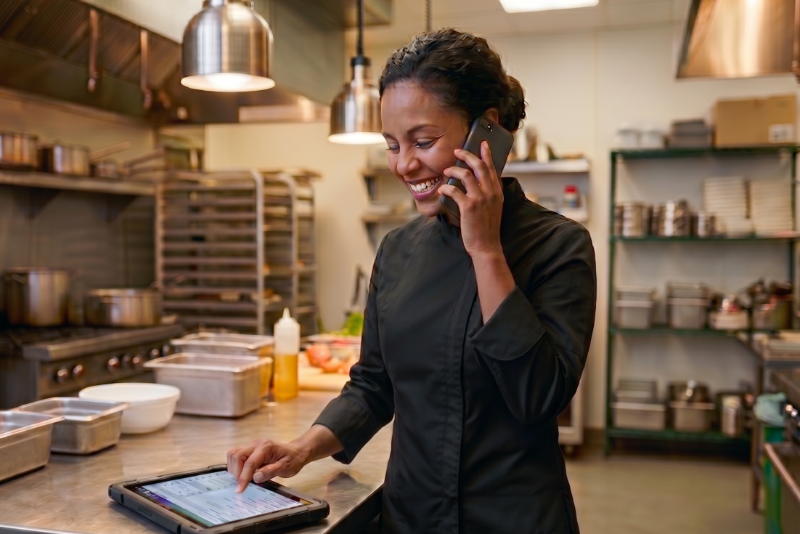 A kitchen staff member in a commercial kitchen talking on a smartphone while using a tablet.